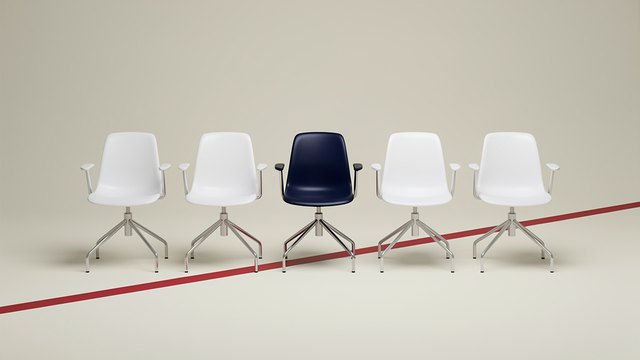 Five white office chairs in a row on a pale studio floor, the middle one navy blue, with a red stripe running diagonally across the floor.